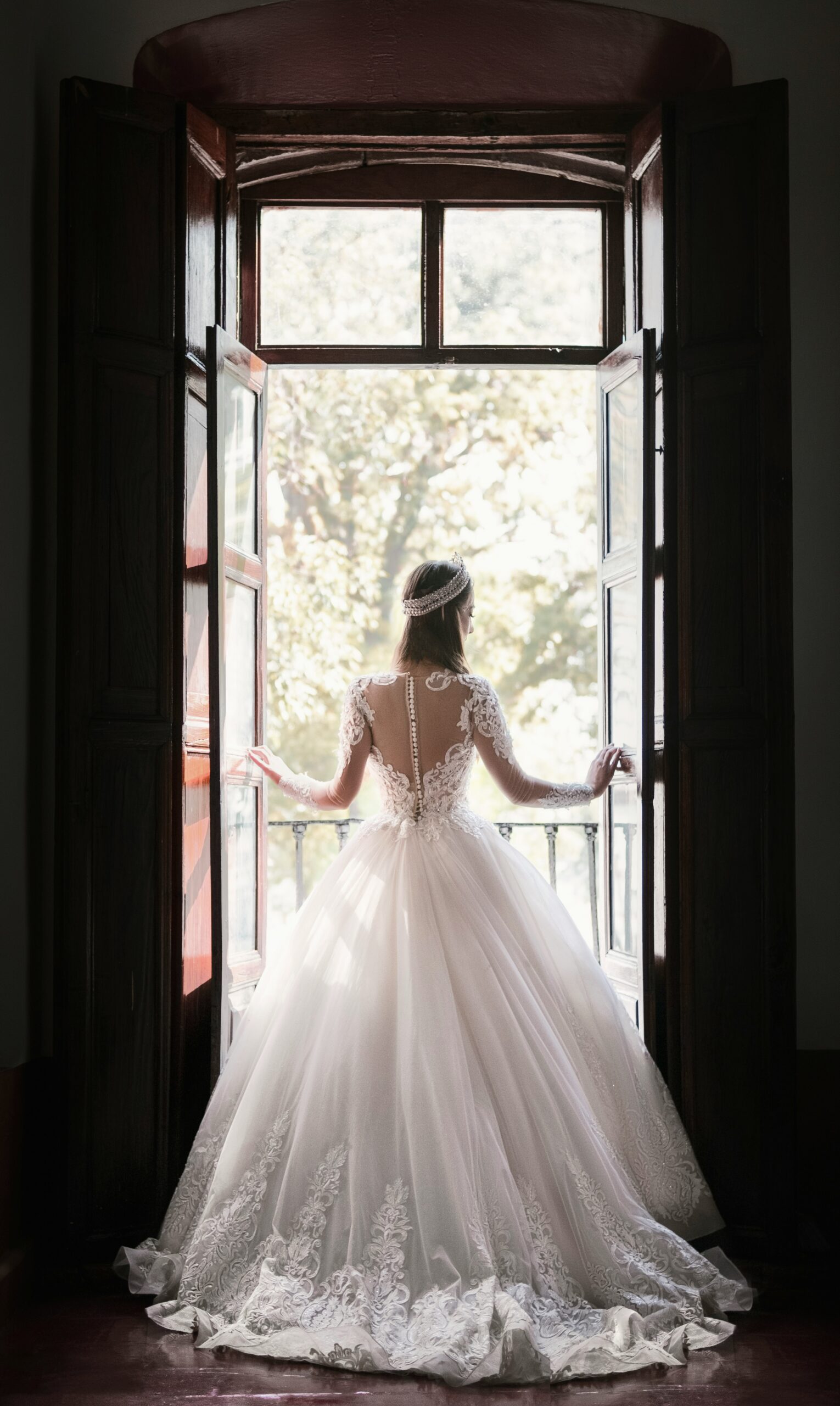 A bride in a lace ball gown and tiara, seen from behind, looking out an open window onto a balcony.