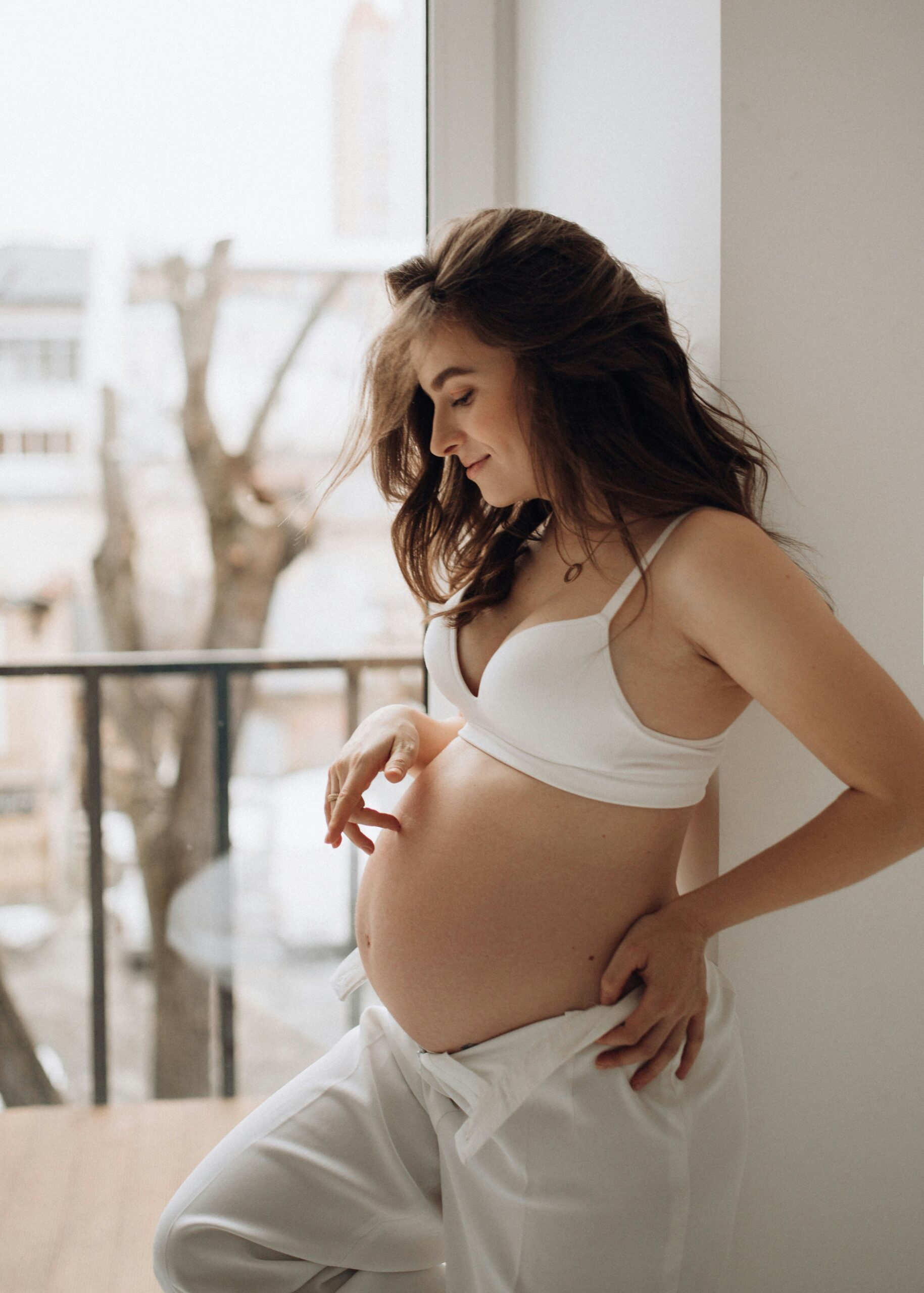 A pregnant woman in a white bra and trousers, touching her belly while leaning against a bright window frame.