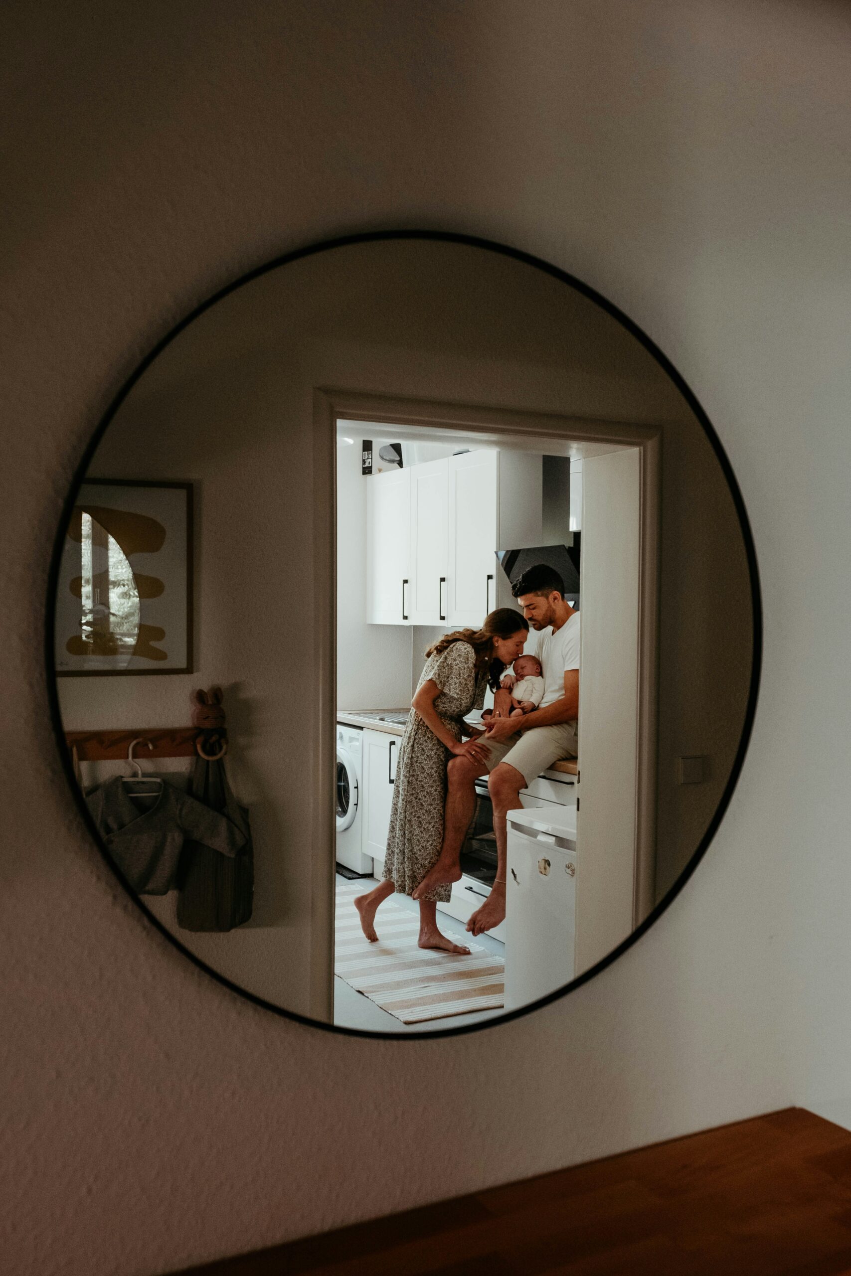 A circular mirror reflecting a couple in a kitchen doorway, the father holding their newborn baby.
