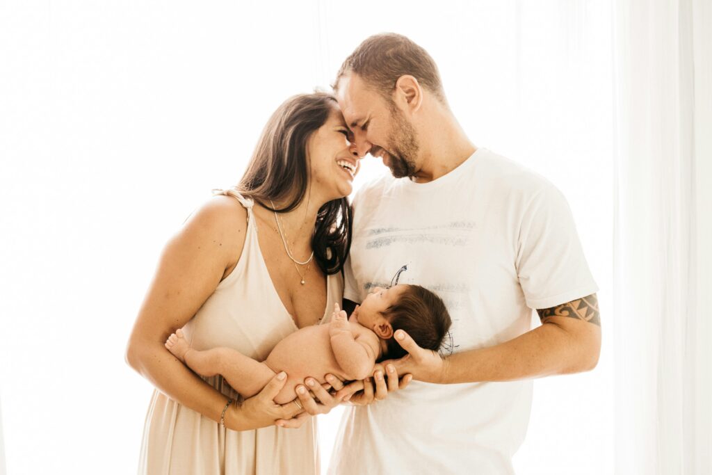 Parents smiling nose-to-nose while holding their newborn baby in soft window light.