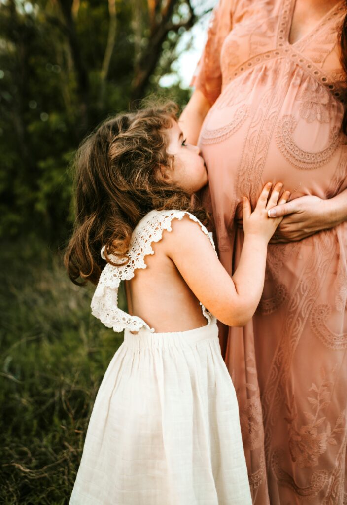A young girl in a white dress kissing her pregnant mother's belly outdoors at golden hour.