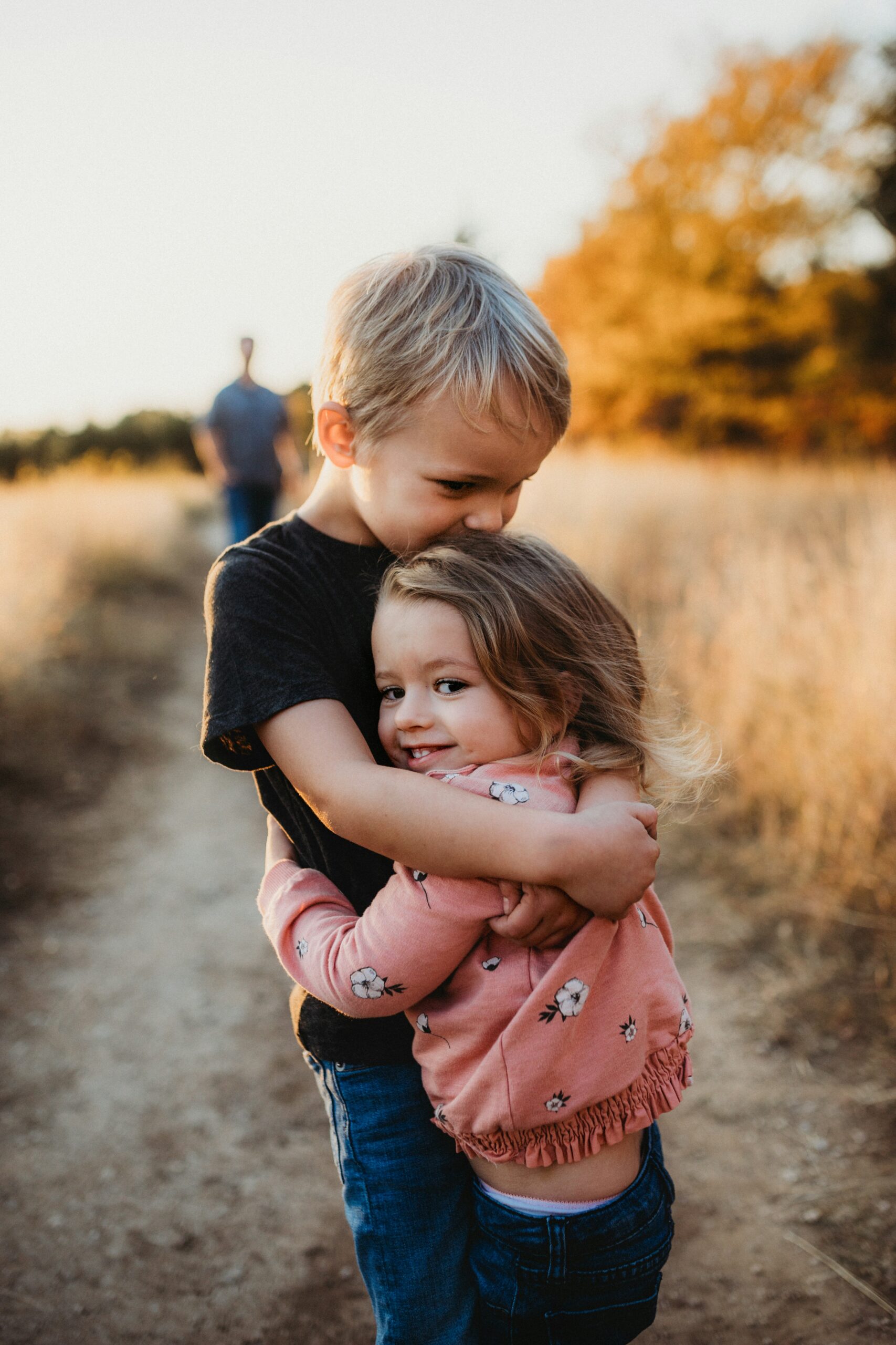 An older brother hugging his younger sister on a dirt path through a golden field at sunset.