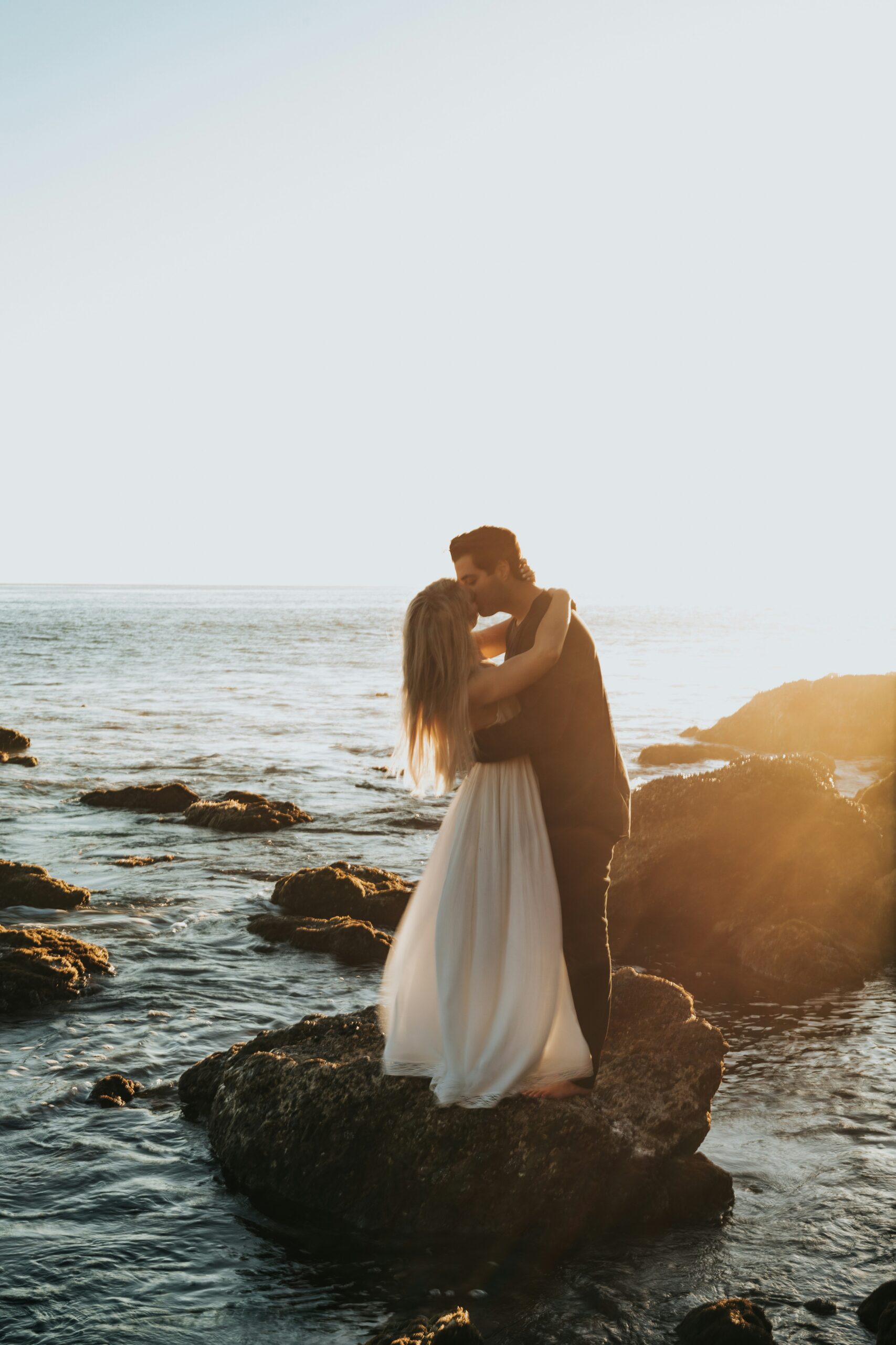 A couple in wedding attire kissing while standing on rocks at the ocean shore during golden hour.
