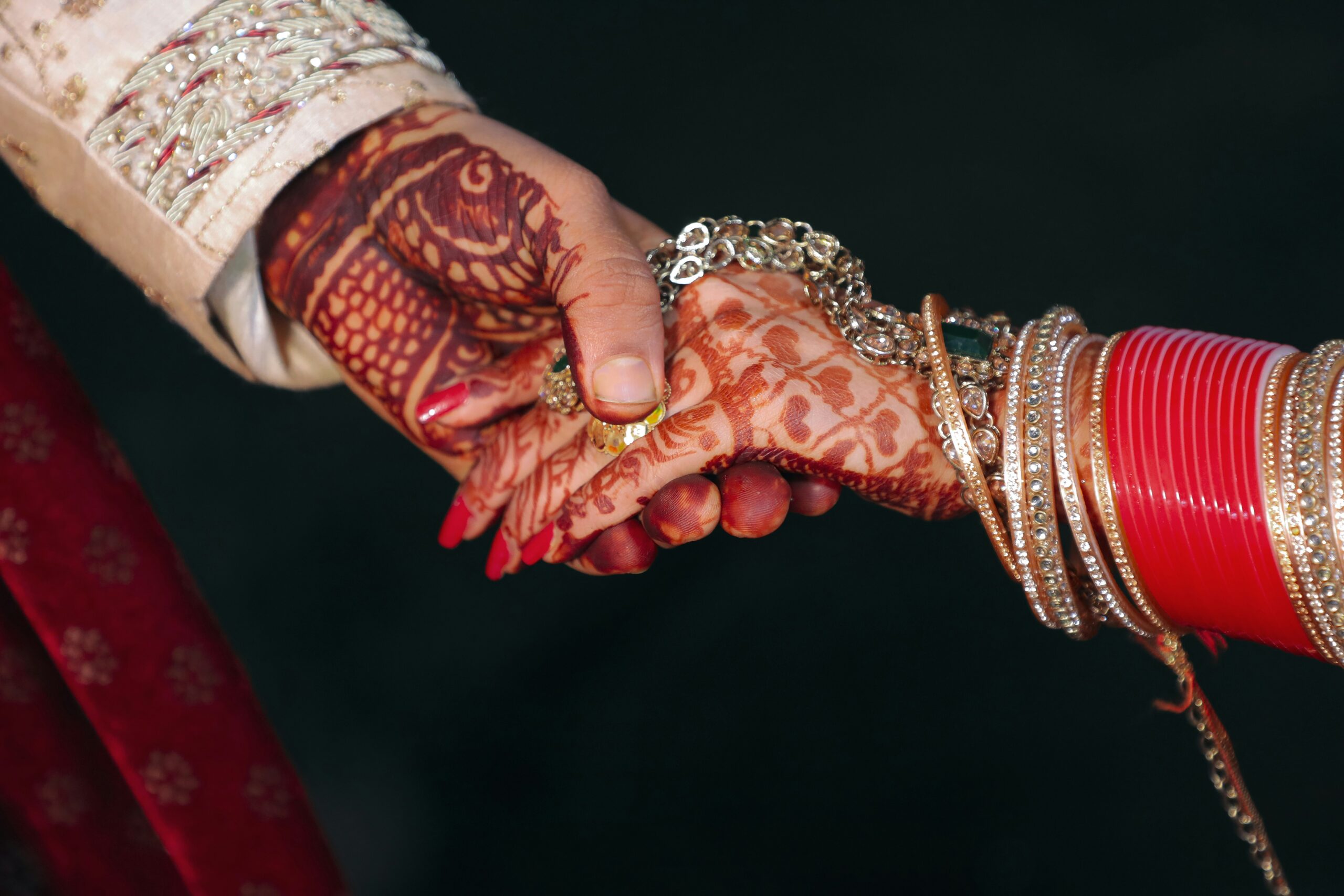 Close-up of a bride and groom holding hands, the bride's hand decorated with intricate henna and bangles.