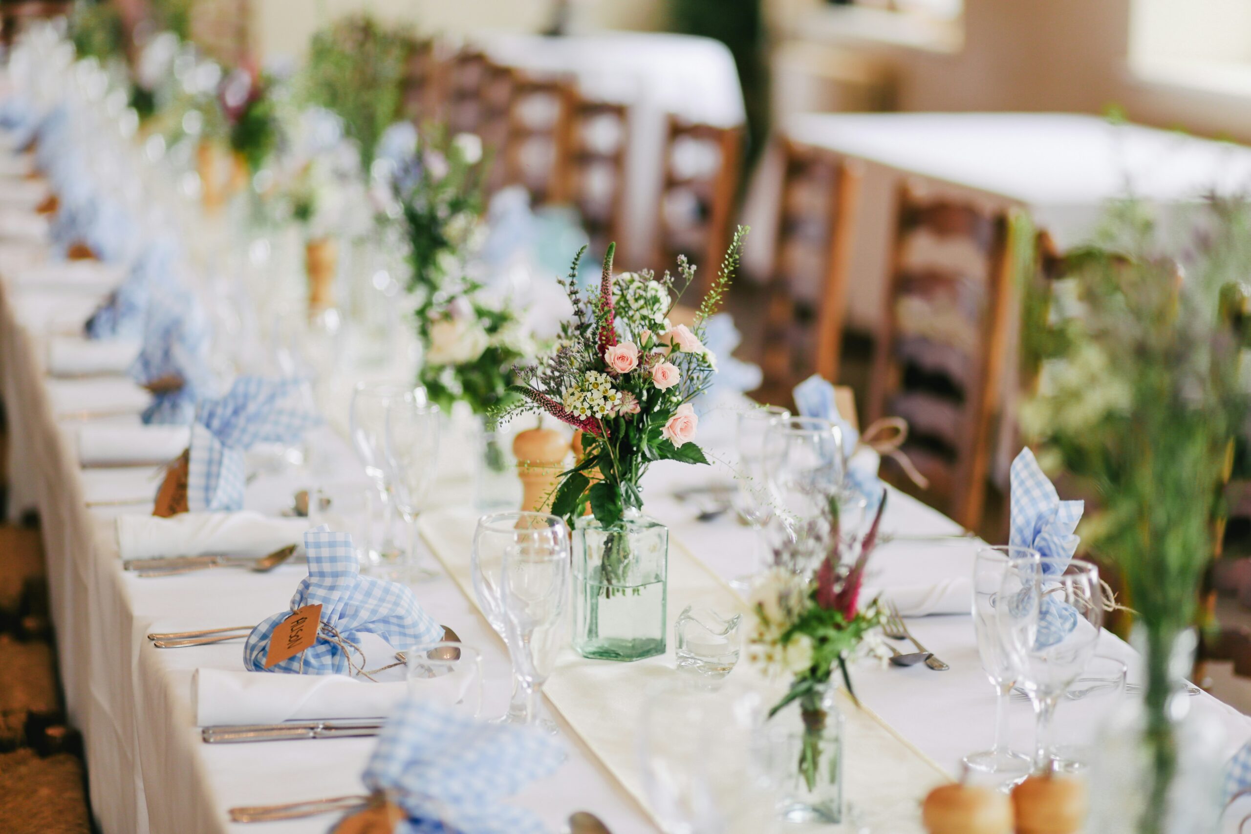 A long wedding reception table set with white linens, wildflower centerpieces, and gingham napkin ties.