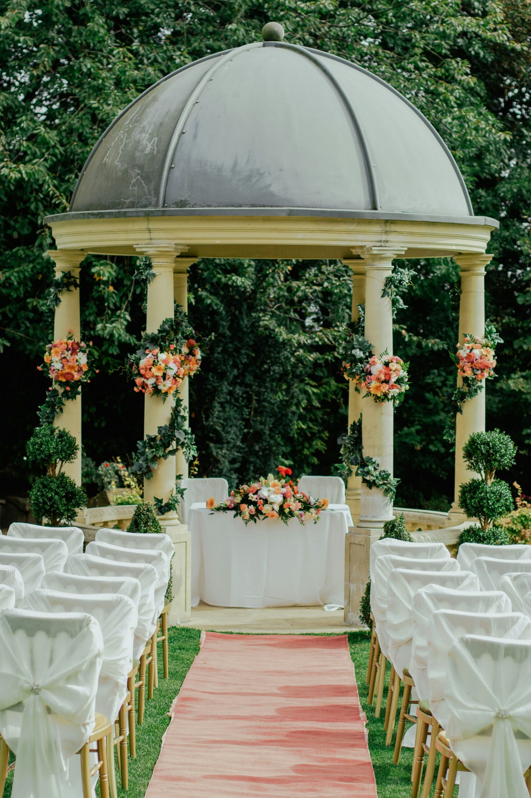 An ornate stone gazebo decorated with flowers, set up for an outdoor wedding ceremony with rows of covered chairs.