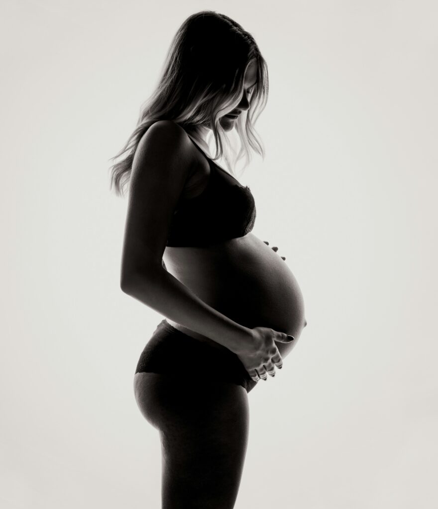 Side-profile silhouette portrait of a pregnant woman in lingerie, hands cradling her belly, high-contrast black and white.
