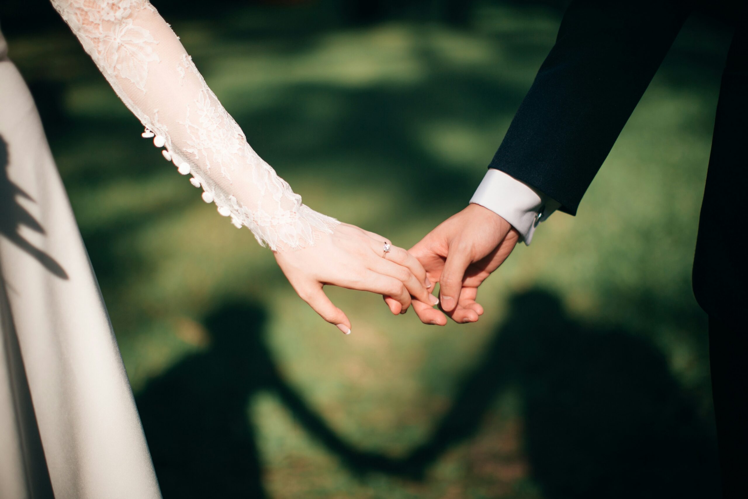 Close-up of a bride and groom holding hands, lace sleeve and engagement ring visible, soft green garden background.